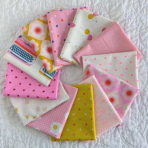 May include: A collection of folded fabric squares arranged in a circular fan pattern. The textiles feature various patterns including pink hearts, yellow and pink flowers, cherries, a blue typewriter, small dots, grid lines, and fried eggs.