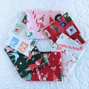 May include: A selection of folded Christmas fabric squares. The fabrics display festive designs such as elves, candy canes, stamps, and snowflakes in red, green, pink, and white. The fabrics are arranged on a white quilted surface.