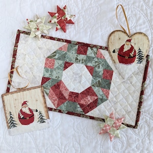 Puede incluir: Un camino de mesa acolchado con un diseño de patchwork rojo y verde que presenta una corona en el centro. El camino de mesa está rodeado de cinco adornos de tela hechos a mano, incluyendo un corazón, estrellas y un adorno de Papá Noel.