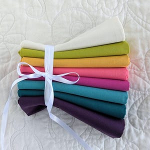May include: A bundle of colourful fabric squares in shades of white, green, yellow, pink, purple, and teal, tied with a white ribbon. The fabric is neatly folded and stacked, creating a visually appealing arrangement.