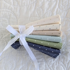 May include: A bundle of six fabric pieces in shades of cream, beige, blue and green, each featuring a polka dot design. The fabric is secured with a white ribbon, creating a tidy stack. The fabrics are displayed on a white, quilted background.