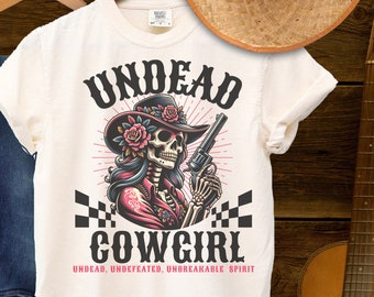 May include: White t-shirt with a graphic of a skeleton cowgirl holding a gun. The text "Undead Cowgirl" is printed in black with the words "Undead, Undefeated, Unbreakable Spirit" printed below. The shirt is on a wooden background with a guitar and a straw hat.