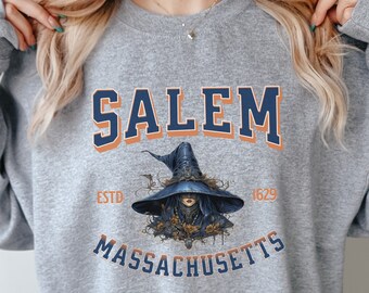 Op de afbeelding: Een grijze sweatshirt met het woord "SALEM" in marineblauw en oranje. Daaronder een illustratie van een heks met een blauwe hoed. De woorden "ESTD 1629" en "MASSACHUSETTS" staan ook op de sweatshirt.