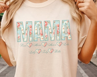May include: A light beige t-shirt with the word "MAMA" in large, floral letters. The letters are surrounded by a light blue border. Below the word "MAMA" are five hearts with the names "Rose", "Missy", "Anya", "Lilly", "Toby", and "Bella" written inside them.