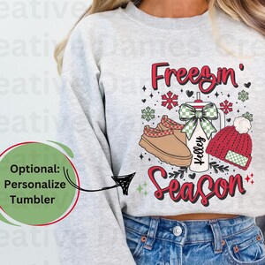 May include: A light gray sweatshirt with a winter-themed graphic. The graphic includes the text "Freezin' Season" in red, along with images of a mug, a hat, and a pair of boots. The sweatshirt is paired with blue jeans.