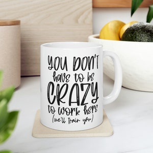 May include: White ceramic mug with the text "YOU DON'T have to be CRAZY to work here (we'll train you)" in black. The mug is on a beige coaster, with a white handle. The background includes a wooden surface and a bowl of fruit.