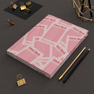 May include: A pink and white journal featuring a repeating pattern of "Guest Check" receipts. The journal is on a dark grey surface, with gold and black pencils and gold binder clips nearby.