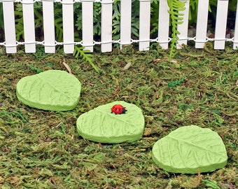 May include: Three green leaf-shaped stepping stones with a red ladybug on the middle stone. The stones are on a bed of green moss.