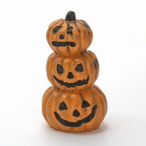 May include: A stack of three orange ceramic jack-o'-lantern pumpkins with black painted faces. The pumpkins are stacked on top of each other, with the smallest pumpkin on top.