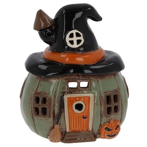 May include: A ceramic pumpkin-shaped house with a glossy green finish, featuring an orange door, brown windows, and a black witch hat roof. The structure includes a small broom leaning against the door and a carved jack-o'-lantern at the base.