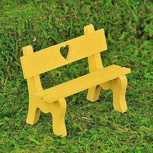 Puede incluir: Un peque&ntilde;o banco de madera amarillo con un recorte en forma de coraz&oacute;n en el respaldo. El banco tiene un asiento de listones y patas curvas. Se encuentra sobre un fondo de musgo verde.