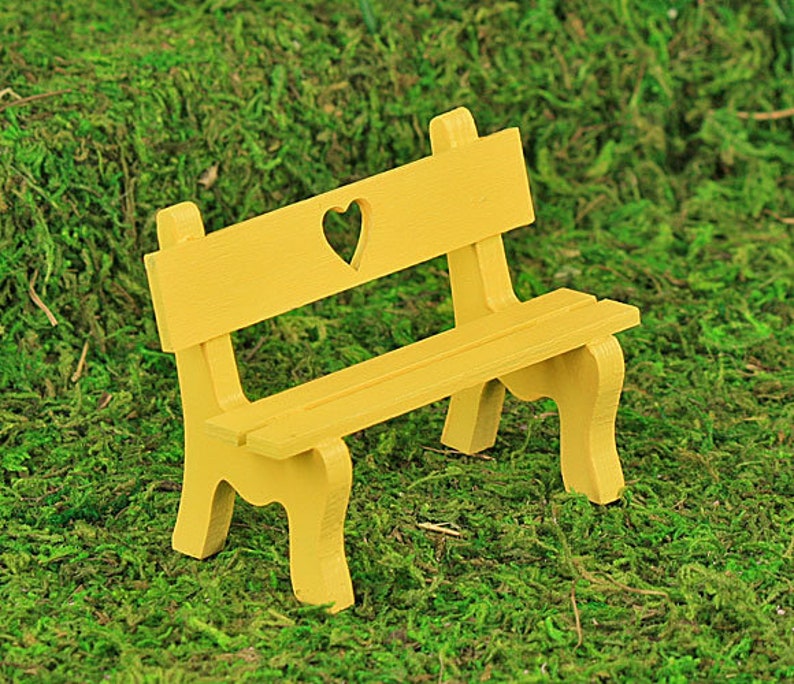 Puede incluir: Un peque&ntilde;o banco de madera amarillo con un recorte en forma de coraz&oacute;n en el respaldo. El banco tiene un asiento de listones y patas curvas. Se encuentra sobre un fondo de musgo verde.
