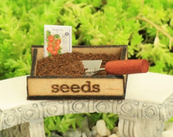 Può includere: Un vassoio per semi in legno in miniatura con un piccolo pacchetto di semi, una paletta di terra marrone e una piccola vanga. Il vassoio ha la scritta "seeds" stampata sul davanti.