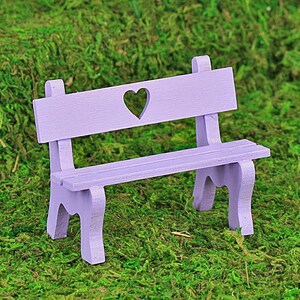 Puede incluir: Un peque&ntilde;o banco de madera color lavanda con un recorte en forma de coraz&oacute;n en el respaldo. El banco tiene un asiento de listones y patas curvas, sobre musgo verde.