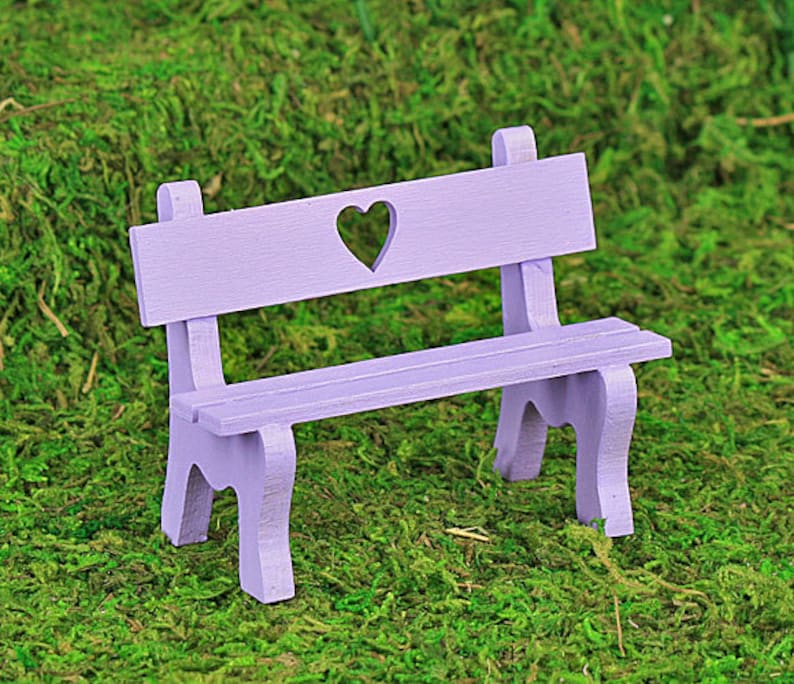 Puede incluir: Un peque&ntilde;o banco de madera color lavanda con un recorte en forma de coraz&oacute;n en el respaldo. El banco tiene un asiento de listones y patas curvas, sobre musgo verde.