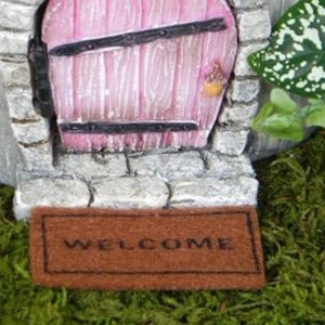 Op de afbeelding: Een miniatuur welkommat met het woord "WELCOME" in bruin op een bruine achtergrond. De mat ligt voor een miniatuur stenen deur met een roze deur en zwarte ijzeren accenten.