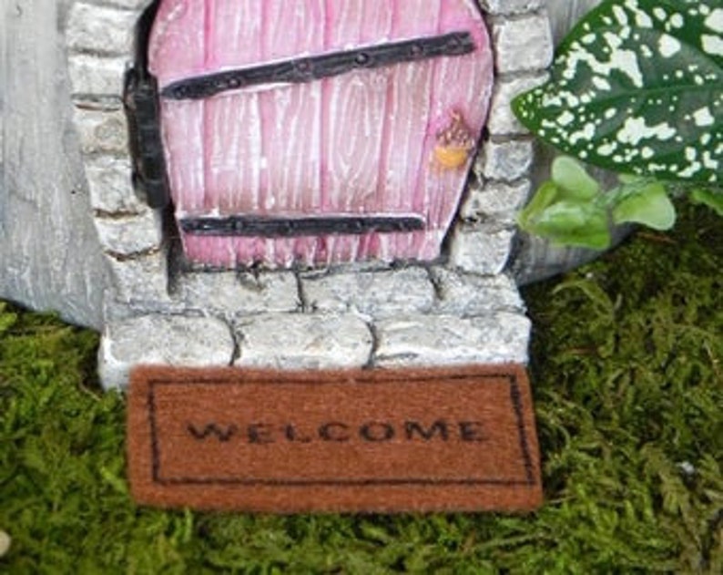 Op de afbeelding: Een miniatuur welkommat met het woord "WELCOME" in bruin op een bruine achtergrond. De mat ligt voor een miniatuur stenen deur met een roze deur en zwarte ijzeren accenten.