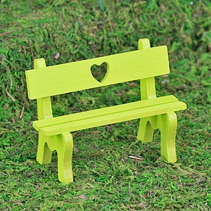 Puede incluir: Un peque&ntilde;o banco de madera verde lima con un recorte en forma de coraz&oacute;n en el respaldo. El banco tiene un asiento de listones y cuatro patas. Se encuentra sobre un fondo de musgo verde, lo que sugiere un jard&iacute;n en miniatura o un entorno de hadas.