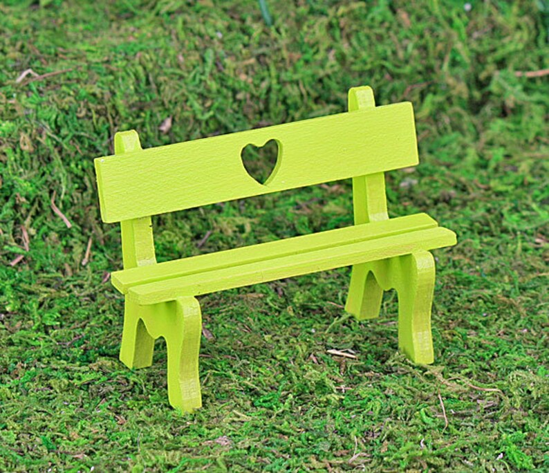 Puede incluir: Un peque&ntilde;o banco de madera verde lima con un recorte en forma de coraz&oacute;n en el respaldo. El banco tiene un asiento de listones y cuatro patas. Se encuentra sobre un fondo de musgo verde, lo que sugiere un jard&iacute;n en miniatura o un entorno de hadas.