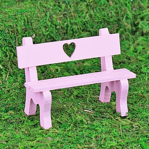 Puede incluir: Un peque&ntilde;o banco de madera rosa claro con un recorte en forma de coraz&oacute;n en el respaldo. El banco tiene un dise&ntilde;o sencillo con cuatro patas y est&aacute; sobre un fondo de musgo verde.
