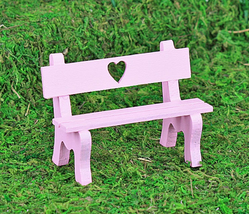 Puede incluir: Un peque&ntilde;o banco de madera rosa claro con un recorte en forma de coraz&oacute;n en el respaldo. El banco tiene un dise&ntilde;o sencillo con cuatro patas y est&aacute; sobre un fondo de musgo verde.