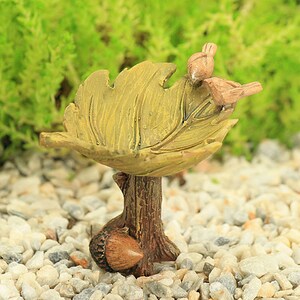 Può includere: Un bagnetto per uccelli in miniatura realizzato in resina, a forma di foglia con una base a tronco d'albero marrone e una ghianda. Due piccoli uccelli sono appollaiati sul bordo della foglia.