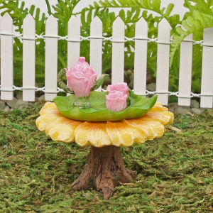 Könnte beinhalten: Ein Miniatur-Tee-Set mit einer rosa Teekanne und Tassen, das auf einem gelben, pilzförmigen Tisch mit einem grünen Blattdesign steht. Das Set ist von grünem Moos umgeben.