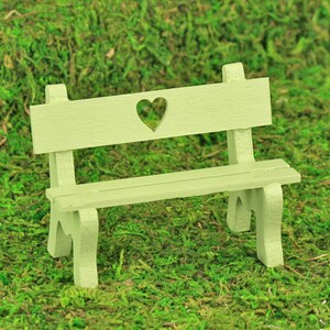 Puede incluir: Un peque&ntilde;o banco de madera verde claro con un recorte en forma de coraz&oacute;n en el respaldo. El banco tiene un asiento de listones y est&aacute; sobre un fondo de musgo verde, lo que sugiere un jard&iacute;n o un entorno al aire libre.