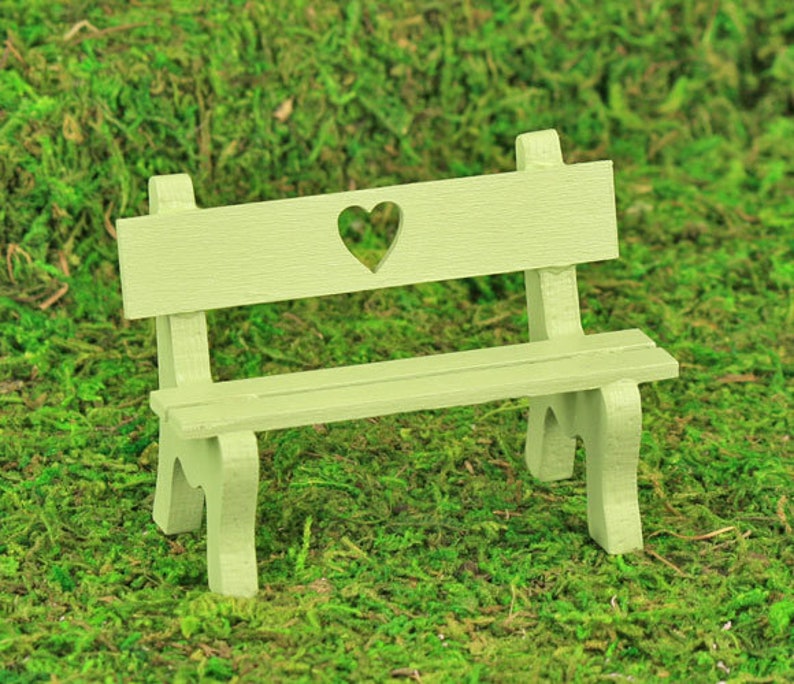 Puede incluir: Un peque&ntilde;o banco de madera verde claro con un recorte en forma de coraz&oacute;n en el respaldo. El banco tiene un asiento de listones y est&aacute; sobre un fondo de musgo verde, lo que sugiere un jard&iacute;n o un entorno al aire libre.