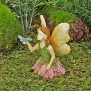 Könnte beinhalten: Eine Miniatur-Feenfigur mit hellem Teint, braunem Haar und gelben Flügeln. Die Fee trägt ein grünes Oberteil und einen rosa Rock und hält einen blauen Schmetterling. Die Figur befindet sich in einer moosigen Gartenszene.