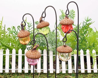 May include: Five miniature acorn-shaped glass ornaments hang from metal stakes. The ornaments are in shades of red, green, yellow, and pink. The stakes are inserted into the ground in front of a white picket fence.