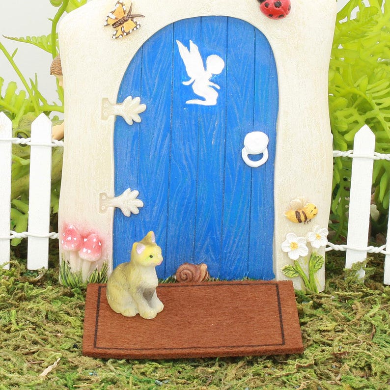 Op de afbeelding: Een miniatuur blauwe deur met een wit silhouet van een fee, een witte deurklink en een bruine deurmat. Een klein grijs en wit kattenbeeldje staat op de mat.