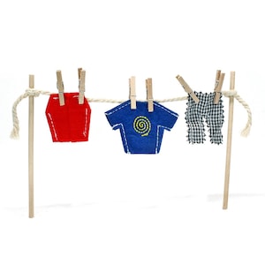Puede incluir: Una cuerda de tender ropa en miniatura con tres prendas colgadas. Una parte superior roja geométrica, una camiseta azul con un diseño de espiral amarilla y pantalones a cuadros blancos y negros están sujetas a la cuerda con pinzas de madera. La cuerda está sostenida por dos postes de madera.