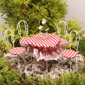 May include: A miniature white patio set with a red and white checkered tablecloth, four chairs, and a table. The set is surrounded by small white rocks and green plants.