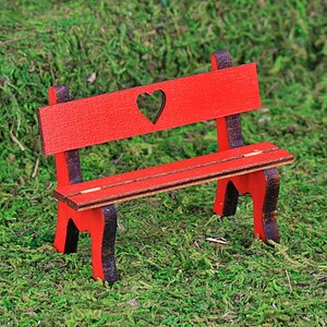 Puede incluir: Un banco de madera rojo en miniatura con un recorte en forma de coraz&oacute;n en el respaldo. El banco tiene detalles en marr&oacute;n oscuro y est&aacute; sobre un fondo de musgo verde. Este banco decorativo es ideal para jardines de hadas o casas de mu&ntilde;ecas.