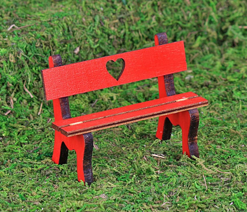Puede incluir: Un banco de madera rojo en miniatura con un recorte en forma de coraz&oacute;n en el respaldo. El banco tiene detalles en marr&oacute;n oscuro y est&aacute; sobre un fondo de musgo verde. Este banco decorativo es ideal para jardines de hadas o casas de mu&ntilde;ecas.