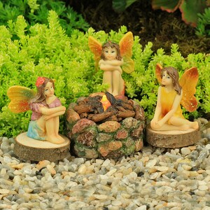 Peut inclure: Trois fées miniatures sont rassemblées autour d'un petit foyer en pierre avec une lumière orange incandescente. Les fées portent des robes colorées et ont des ailes. Le foyer est fait de petites pierres et a un tas de bûches au centre.