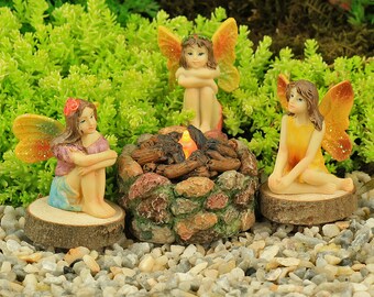 Può includere: Tre fate in miniatura sono riunite intorno a un piccolo focolare di pietra con una luce arancione incandescente. Le fate indossano abiti colorati e hanno le ali. Il focolare è fatto di piccole pietre e ha un mucchio di ceppi al centro.