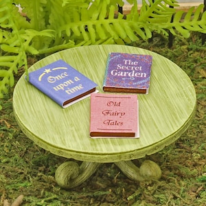 Peut inclure: Trois livres miniatures sur une table en bois vert. Les livres sont intitulés "Il était une fois", "Le jardin secret" et "Les vieux contes de fées".