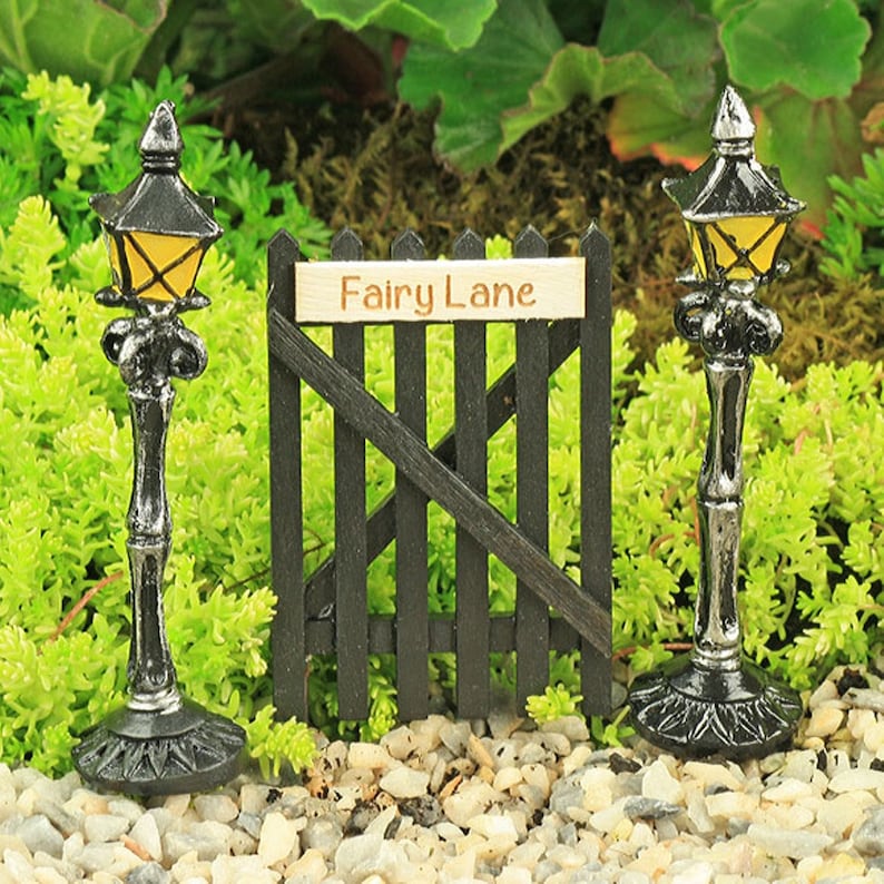 May include: Two miniature black lamp posts with yellow lights and a black wooden gate with a sign that reads "Fairy Lane".