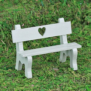Puede incluir: Un banco en miniatura de color gris claro con un recorte en forma de coraz&oacute;n en el respaldo. El banco tiene un asiento de listones y patas curvas, dise&ntilde;ado para una casa de mu&ntilde;ecas o un jard&iacute;n en miniatura. El banco est&aacute; sobre musgo verde.