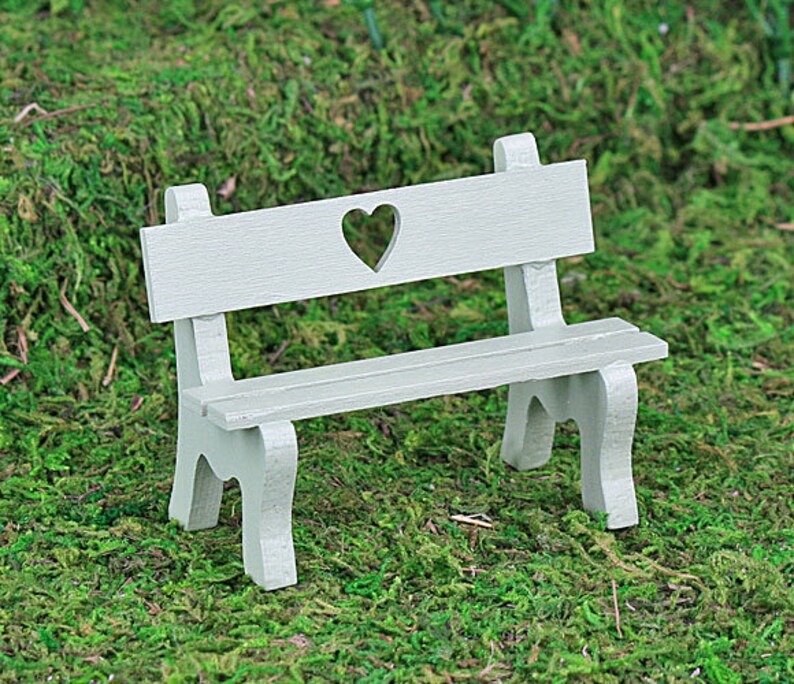 Puede incluir: Un banco en miniatura de color gris claro con un recorte en forma de coraz&oacute;n en el respaldo. El banco tiene un asiento de listones y patas curvas, dise&ntilde;ado para una casa de mu&ntilde;ecas o un jard&iacute;n en miniatura. El banco est&aacute; sobre musgo verde.