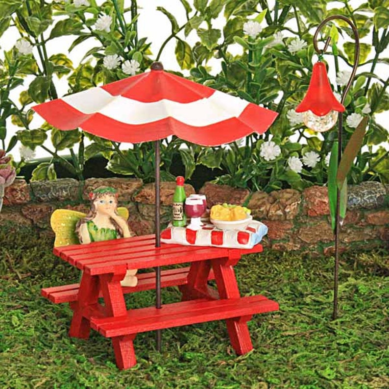 Puede incluir: Una mesa de picnic roja en miniatura con una sombrilla a rayas rojas y blancas, una figurita de hada sentada en un banco y una botella de vino y una copa de vino en la mesa.