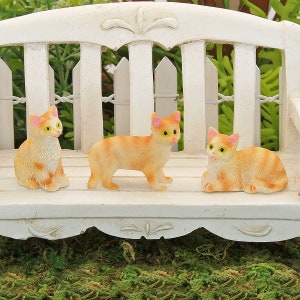 May include: Three miniature orange tabby cats sitting on a white bench. The cats are made of plastic and have realistic details.