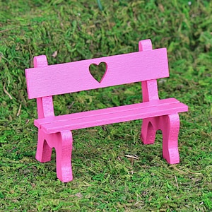 Puede incluir: Un peque&ntilde;o banco de madera rosa con un recorte en forma de coraz&oacute;n en el respaldo. El banco tiene un asiento de listones y est&aacute; sobre un lecho de musgo verde. El banco es un objeto decorativo.