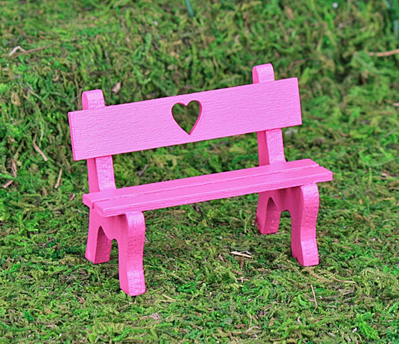Puede incluir: Un peque&ntilde;o banco de madera rosa con un recorte en forma de coraz&oacute;n en el respaldo. El banco tiene un asiento de listones y est&aacute; sobre un lecho de musgo verde. El banco es un objeto decorativo.