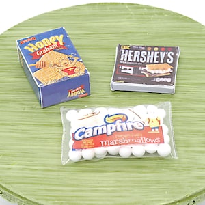 Könnte beinhalten: Nahaufnahme von drei Schachteln mit Zutaten für die Zubereitung von S'mores. Die Schachteln befinden sich auf einer grünen Oberfläche. Die Schachteln sind mit "Honey Graham", "Hershey's Milk Chocolate" und "Campfire Marshmallows" beschriftet.