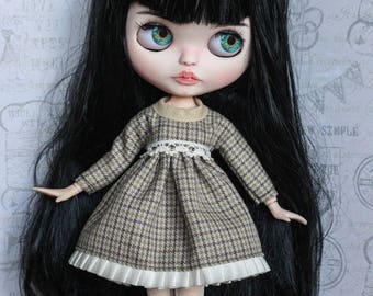 May include: A Blythe doll with long black hair and large, teal eyes. The doll is dressed in a brown and white plaid dress with a white lace collar and trim. The doll wears white boots with white fur trim. The text "Soul Blythe" is in the upper right corner.