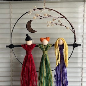 Puede incluir: Un colgante de pared caprichoso con tres muñecas de hilo en rojo, verde y morado, cada una con una cabeza de madera y un peinado único. Las muñecas están suspendidas de un aro de metal negro adornado con una luna creciente y detalles florales.