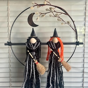 May include: A Halloween-themed wall hanging featuring two yarn witches with black and silver dresses, black hats, and broomsticks. The witches are suspended within a black metal circle, with a crescent moon and decorative branches above.
